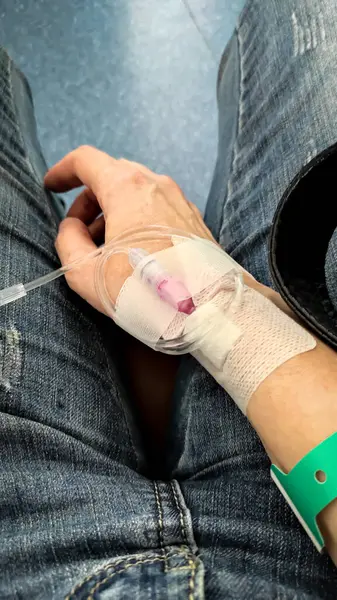 Klinik ortamda damar içi kateteri olan bir elin yakın çekimi. Kot pantolonlu bir hasta hastane bileziğiyle hastanede oturuyor..