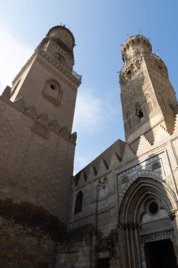 Mısır 'ın Kahire kentindeki Al-Azhar Camii' nin alçak açılı görüntüsü. Resim, caminin karmaşık taş işçiliğini, dekoratif kemerlerini ve minarelerini gözler önüne seriyor. Duvarlardaki ve kulelerdeki detaylı oymalar ve desenler caminin tarihi eserinin bir göstergesi.