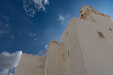 Geleneksel Arap şapkası ve beyaz giysisi giyen Müslüman bir adam tarihi bir cami veya benzer bir binanın önünde görülüyor. Görüntü binanın mimarisini gösteriyor, açık bej / bronz renkli dış görünüşü ve kısmen görünür bir m