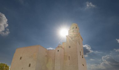 Resmin odak noktası, muhtemelen çöl ikliminde bulunan büyük, açık bej bir cami. Güneş gökyüzünde yüksekte, gölgeler bırakıyor ve minareden yayılan güneş ışınları yaratıyor. Geleneksel Müslüman giysileri içinde çok sayıda insan.