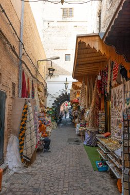 Fas 'ta (eski şehir) dar bir ara sokak canlı tezgahlarla doludur. Halılar, sepetler, mücevherler ve hediyelik eşyalar da dahil olmak üzere çeşitli satıcılar (tanımlanamayan) mallarını sergiliyor ve satıyor. Geleneksel Fas kıyafetleri içinde bir adam tezgahta oturuyor.