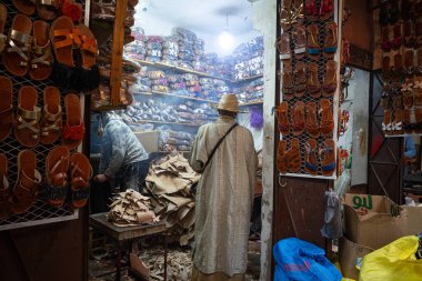 Hareketli bir deri sandalet dükkanı. İşçiler (tanımlanamayan) ön plandaki bir masanın üzerindeki hammadde yığınları gibi görünen deri derileri işliyorlar. Geleneksel açık bej bornoz ve hasır şapkalı bir adam muhtemelen bir müşteri ya da dükkan sahibidir.