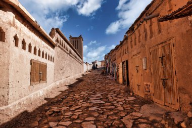 Dar, güneşli bir ara sokak, muhtemelen Fas 'ta bulunan geleneksel bir Berber köyünden geçiyor. Bazı bireyler (tanımlanamayan), muhtemelen dükkan sahipleri veya yoldan geçenler, düz olmayan, taşlarla döşenmiş cadde boyunca ilerliyorlar. Bu binalar...