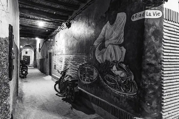 Fas 'ın bir şehrinde dar bir ara sokak, muhtemelen Marakeş' te gösteriliyor. Duvarın yanında bir motosiklet park edilmiş. Duvar canlı bir duvar resmiyle süslenmiş ve bir su kütlesi tarafından yapılmış, geleneksel giysiler içinde bir figür resmedilmiş. Duvar resmi...