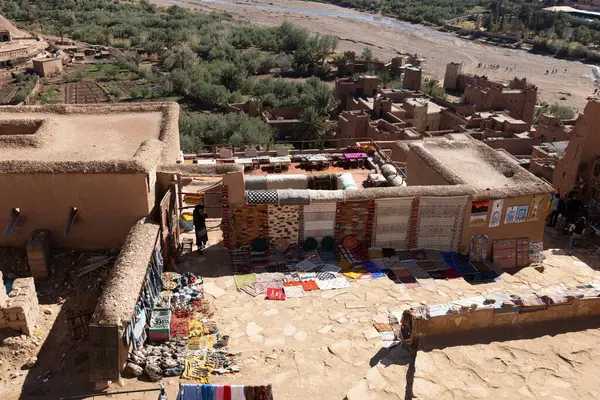 Geleneksel bir Kuzey Afrika kasabasında hareketli bir pazar sahnesi. Yerde ve duvarlar boyunca çeşitli renkli halılar ve diğer el yapımı mallar satışa sunuluyor. Çeşitli turist ve yerel tedarikçiler bulunurken, bazı satıcılar fuara katılıyor