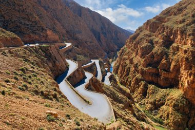 Derin bir kanyondan geçen kıvrımlı bir dağ yolu yılanı, muhtemelen Fas 'ın Atlas Dağları' nda. Yollarda çok sayıda araç (en az iki araba ve bir minibüs), dik ve dar geçitte yol alırken görülüyor. Sert, terasotta renkli kayalıklar ve... 