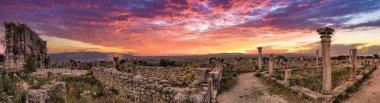 Turistler antik Roma şehri Volubilis, Fas 'ın kalıntılarını keşfediyorlar. Görüntü, iyi korunmuş taş duvar ve kemerlerin bir bölümünü ve bir zamanlar büyük yapıların kalıntıları arasında turistlerin (muhtemelen turistler) yürüdüğü bir bölümü gösteriyor. Bulutlu 