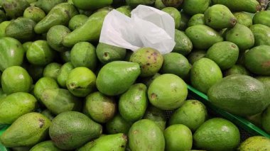 Olgun yeşil avokado yığınları süpermarkette bir sepetin üzerinde duruyor, bazıları plastikle kaplı. Pürüzsüz, parlak derileri ve taze dokuları görülebilir. Yemek ve market görselleri için mükemmel.