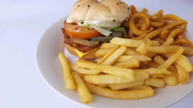 Normal ve kıvırcık patatesli sulu bir hamburgerin dönen tabağı, temiz beyaz arka plana karşı 45 derecelik açıyla vurulmuş. Çıtır patatesler ve mükemmel istiflenmiş bir hamburger ağız sulandırıcı bir manzara yaratır.