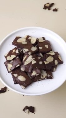 Dönen bir tabak bademli brownie beyaz bir plaka üzerinde inceleniyor, bej arka plan, 45 derecelik açıyla çekiliyor. Tatlı, atıştırmalık ve yemek sunumu için mükemmel bir dikey video..