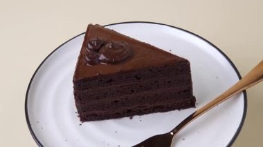 Zengin bir çikolatalı pasta siyah kenarlı beyaz bir tabakta döner, yanında da bir gül altın çatalı vardır. Bej arka plan zarafeti artırır. Tatlı ve fırın görselleri için mükemmel dikey stok videosu.