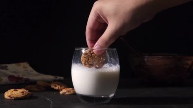 Süte batırılmış kurabiyenin yakın plan çekimi, el modeli kurabiyeyi yukarıda tutuyor, süt yavaşça bardağa geri damlıyor. Kurabiye, peçete ve çikolata kaseli siyah arkaplan..