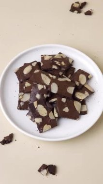 Bademli browni inceltilmiş beyaz bir tabakta, bej arka planda. Bir el modeli 45 derecelik açıyla tek bir zayıf nokta yakalar. Dikey çekim, tatlı ve atıştırmalık için mükemmel..