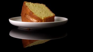 Beyaz tabaktaki sünger kek parlak siyah yansıtıcı yüzeyde dönüyor. Ayna yansıması pastanın altın dokusunu ve yumuşak katmanlarını vurguluyor