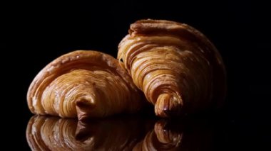 Parlak siyah yansıtıcı yüzeyde dönen üç altın kruvasan, belirgin ayna yansıması, koyu arka plan renklendirme dokusu ve pullu katmanlar. Ay çöreği..