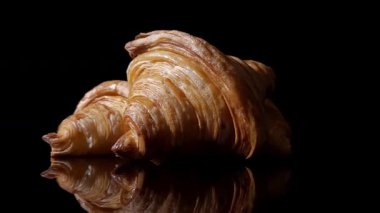 Siyah yansıtıcı yüzeyde saat yönünün tersine dönen üç kruvasan, güçlü ayna yansıması, altın pullu hamur işi dokusunu vurgulayan koyu bir arka plan. Ay çöreği..