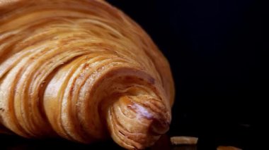 Çerçevenin sol tarafında parlak, siyah yansıtıcı bir yüzey üzerinde dönen bir kruvasanın yakın çekimi. Zengin doku ve altın katmanlar koyu arkaplanla tezat oluşturuyor. Ay çöreği..