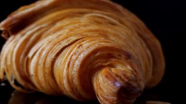 Siyah yansıtıcı arka planda dönen bir kruvasanın yakın plan görüntüsü, çerçevede ortalanmış. Zengin doku, altın katmanlar ve koyu zarif bir arkaplanda güçlü bir yansıma..