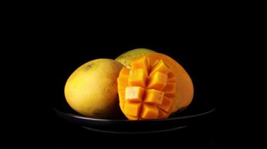 Üç mango siyah plaka üzerinde dönüyor, biri dilimlenmiş kirpi stili, koyu siyah arka planlı yansıtıcı yüzeye odaklı.