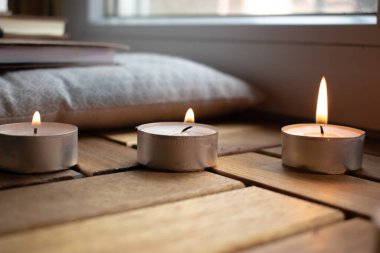 Mumlar ahşap bir yüzey üzerinde yanıyor, sıcak bir ortam yaratıyorlar. Yakınlarda, eskiz defterleri ve desenli bir yastığın üzerinde bir kalem istirahatı. Mum ışığı sahneyi hafifçe aydınlatıyor, samimi ve yaratıcı bir atmosfer izlenimi veriyor.. 