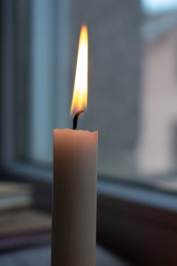 Pencerenin yanında sabit bir alevi olan bir mum. Mum krem renkli ve hafif kıvrımlı fitili var. Bulanık arkaplanda, bir eskiz defteri veya muhtemelen kitaplar dizilmiş olarak görülebilir