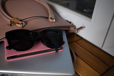 Laptop, pembe defter ve tahta yüzeyde güneş gözlüğü. Yanında altın aksanlı bej bir çanta. Moda, teknoloji ve verimliliği harmanlayan parlak, şık bir dekor..