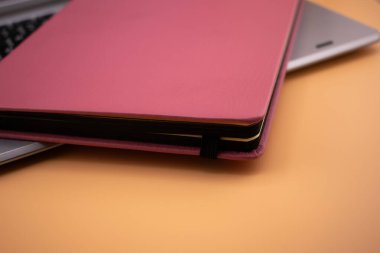 Pembe bir not defteri gümüş bir dizüstü bilgisayarın üzerinde duruyor. Not defteri ve dizüstü bilgisayar kompakt ve düzenli bir uzaktan çalışma ortamı olduğunu gösteriyor. Ögelerin kombinasyonu, pratik ve stil arasındaki dengeyi vurgular..