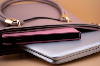 Dizüstü bilgisayar ve defter kısmen leylak rengi bir çantaya sıkıştırılmış. Çantanın altın renginde donanım aksanı var. Şık ve profesyonel bir görünüm sağlıyor. Kurulum açık kahverengi bir yüzeye dayanıyor. Bu da yaratıcı ve düzenli bir çalışma alanına işaret ediyor..