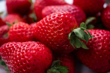 Taze çilek, zengin kırmızı, beyaz bir tabağı dolduruyor, doğal ışıkla vurgulanıyor, yeşil yapraklı üst kontrastla canlı meyveler. Yüksek C vitamini içeriğiyle bilinir