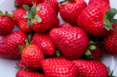 Taze çilek, zengin kırmızı, beyaz bir tabağı dolduruyor, doğal ışıkla vurgulanıyor, yeşil yapraklı üst kontrastla canlı meyveler. Yüksek C vitamini içeriğiyle bilinir