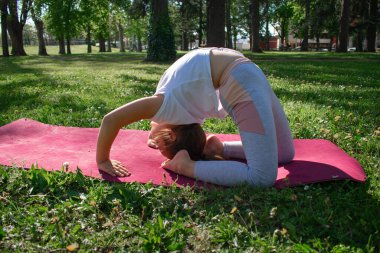 Zarif genç kız, güneşli bir parktaki kestane rengi bir hasırda (Urdhva Dhanurasana varyasyonu) ters köşe yapar. Doğal ortamda esneklik, güç ve bilinçli hareket gösterir. Yoga ve sağlık kavramları için idealdir.