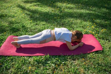 Kararlı genç kız, parlak bir parkta yoga minderinin üzerinde tahta bir duruş sergiliyor. Çekirdek gücü, odaklanmayı ve doğadaki sağlıklı aktiviteyi vurgular. Zindelik ve refah temaları için ideal.