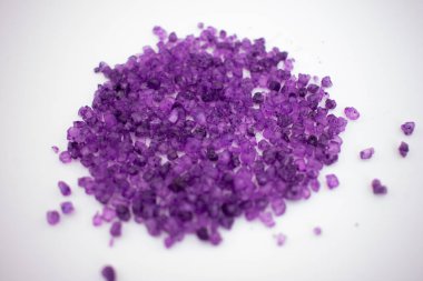 Temiz beyaz bir yüzey üzerinde canlı bir mor banyo tuzu yığını. Rahatlama, aromaterapi ve lüks bir spa deneyimini çağrıştırıyor.
