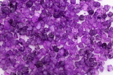 Temiz beyaz bir yüzey üzerinde canlı bir mor banyo tuzu yığını. Rahatlama, aromaterapi ve lüks bir spa deneyimini çağrıştırıyor.