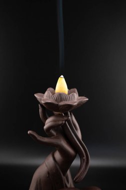 Nilüfer tutan el şeklinde koyu seramik bir tütsü yakıcı ve hafif bir tütsü külahıyla narin bir duman çıkaran sakin bir sahne. Barışı ve aromaterapiyi çağrıştırıyor.