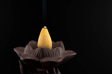 Nilüfer tutan el şeklinde koyu seramik bir tütsü yakıcı ve hafif bir tütsü külahıyla narin bir duman çıkaran sakin bir sahne. Barışı ve aromaterapiyi çağrıştırıyor.