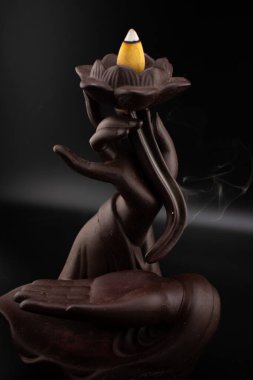 Nilüfer tutan el şeklinde koyu seramik bir tütsü yakıcı ve hafif bir tütsü külahıyla narin bir duman çıkaran sakin bir sahne. Barışı ve aromaterapiyi çağrıştırıyor.