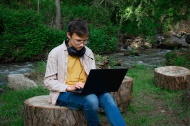 Bu görüntü, kulaklıklı bir gencin dışarıda akan bir derenin yanında dizüstü bilgisayar kullanmasını gösteriyor. Uzak çalışma, çevrimiçi öğrenme ve doğanın teknolojiyle harmanlanması için ideal temalar