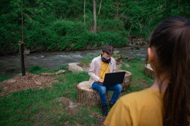 Bu görüntü, dışarıda dizüstü bilgisayarda çalışan bir gencin uzaktan öğrenme ve teknolojiyi doğayla harmanlama teması için ön planda kısmen görünür bir kişi ile birlikte çalıştığını gösteriyor.