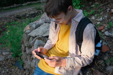 Bu görüntü, harabelerin yanındaki eski bir duvara yaslanırken akıllı telefon kullanan bir genci gösteriyor. Tarihsel ortamlarda, gençlik kültüründe ve gelişigüzel keşiflerde teknoloji temaları için ideal