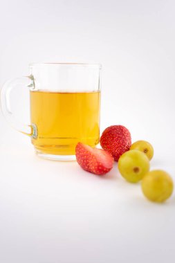 Taze çilek ve yeşil üzüm taşıyan ahşap bir bardak altının yanında altın bitkisel çay bulunan cam bardak. Düzenleme, zarif bir sunumla sağlık ve beslenme üzerine odaklanılmasını öneriyor..