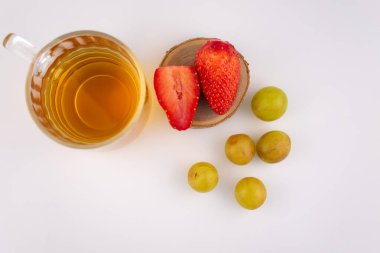 Taze çilek ve yeşil üzüm taşıyan ahşap bir bardak altının yanında altın bitkisel çay bulunan cam bardak. Düzenleme, zarif bir sunumla sağlık ve beslenme üzerine odaklanılmasını öneriyor..