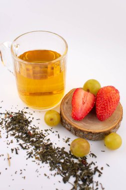 Taze çilek ve yeşil üzüm taşıyan ahşap bir bardak altının yanında altın bitkisel çay bulunan cam bardak. Düzenleme, zarif bir sunumla sağlık ve beslenme üzerine odaklanılmasını öneriyor..