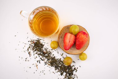 Taze çilek ve yeşil üzüm taşıyan ahşap bir bardak altının yanında altın bitkisel çay bulunan cam bardak. Düzenleme, zarif bir sunumla sağlık ve beslenme üzerine odaklanılmasını öneriyor..