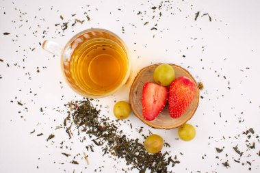 Taze çilek ve yeşil üzüm taşıyan ahşap bir bardak altının yanında altın bitkisel çay bulunan cam bardak. Düzenleme, zarif bir sunumla sağlık ve beslenme üzerine odaklanılmasını öneriyor..