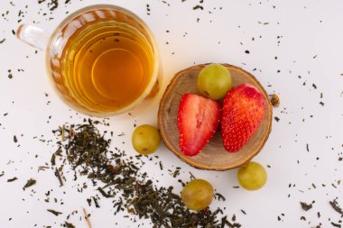 Taze çilek ve yeşil üzüm taşıyan ahşap bir bardak altının yanında altın bitkisel çay bulunan cam bardak. Düzenleme, zarif bir sunumla sağlık ve beslenme üzerine odaklanılmasını öneriyor..