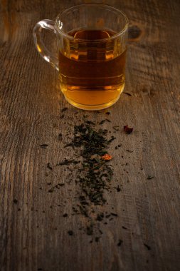 Sadelikte sükuneti bul! Dağılmış çay yaprakları ile kırsal bir ahşap yüzeye odaklanmış cam bardak dolusu yeşil çay, sakinlik ve doğal saflığı çağrıştırıyor. Sağlık, dikkatli yaşam ya da içecek temaları için idealdir.