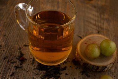 Bu sanatsal kompozisyonda bir bardak yeşil çay, gevşek çay yaprakları ve kırsal ahşap üzerinde taze yeşil üzümler bulunur. Bu da doğal bir refah ve hoş bir basitlik hissi uyandırır. Sağlık blogları, bilinçli yaşam ya da içecek endüstrisi içeriği için ideal