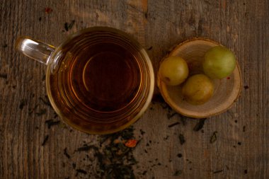 Bu sanatsal kompozisyonda bir bardak yeşil çay, gevşek çay yaprakları ve kırsal ahşap üzerinde taze yeşil üzümler bulunur. Bu da doğal bir refah ve hoş bir basitlik hissi uyandırır. Sağlık blogları, bilinçli yaşam ya da içecek endüstrisi içeriği için ideal