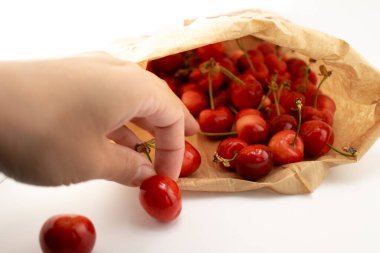Yakın plan çekimde, taze, olgun bir kiraz seçmek için bir kese kağıdına uzanan bir el yakalıyor ve doğal bir iyilik hissi uyandırıyor. Organik gıda, çiftçi pazarı veya çevre dostu yaşam tarzı içeriği için ideal